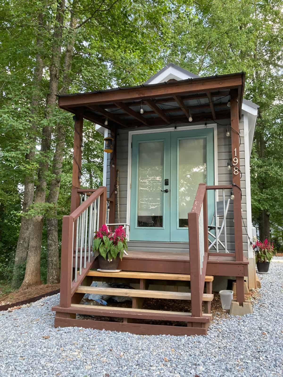 Covered front porch of the Mustard Seed Sprout tiny house with aqua French doors, string lights, and house number 198