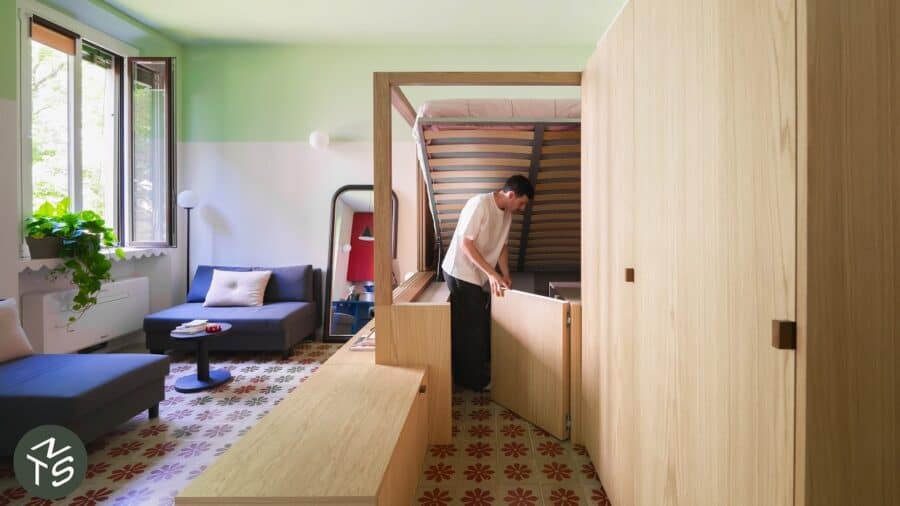 Multifunctional light-oak sleeping alcove with lift-up bed storage, tall wardrobe, navy seating, and patterned Milanese tile floor