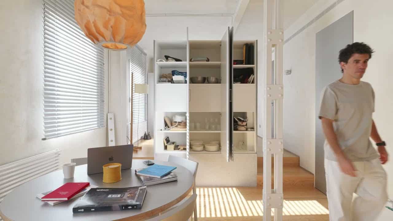 Round dining and work table with a laptop and books under a crumpled paper pendant; freestanding storage unit open to shelves; two wood steps rising beside a white steel column