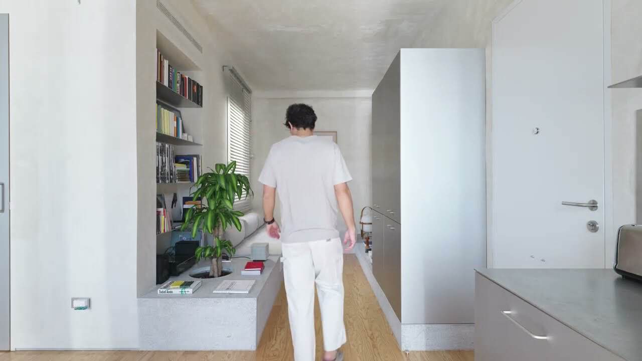 Man walking away down Casa VA’s living run: granite platform with a circular planter and books, recessed wall shelves, freestanding grey storage unit, stainless kitchen counter at the right edge