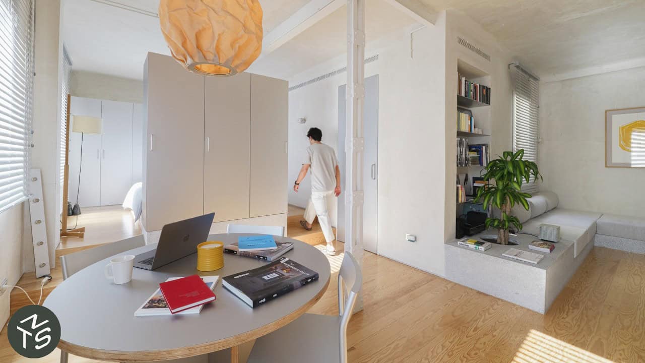 Open-plan Casa VA from the dining end: round work table under a paper pendant, freestanding storage unit, granite platform sofa with a built-in planter, man walking a two-step rise
