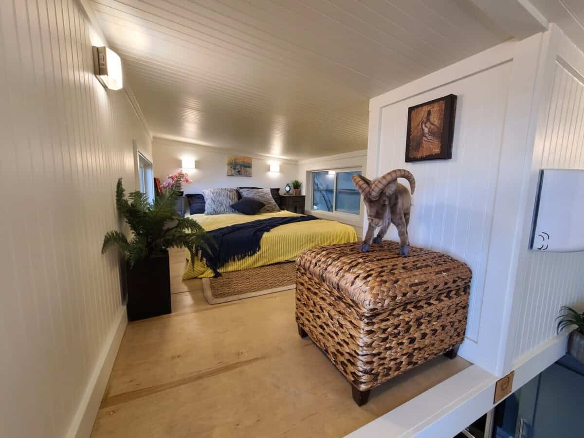 Primary loft with a bed at the far end under a low white ceiling, seen from the top of the stairs