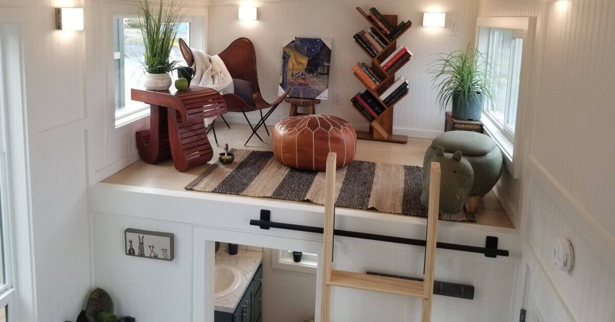Secondary loft staged as a reading area with a leather chair, a pouf, and a bookshelf, above the bathroom with a wood ladder