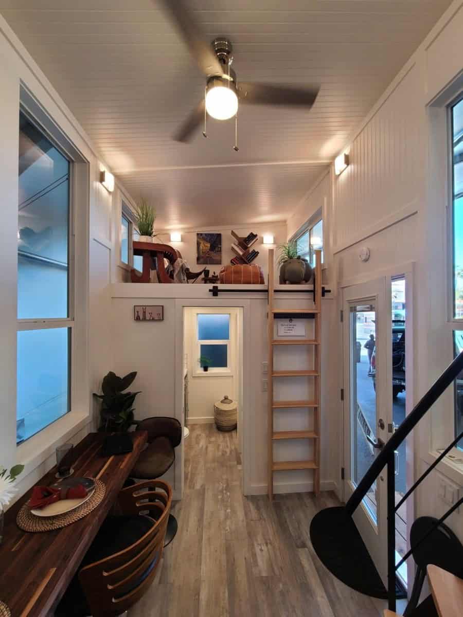 Interior view toward the bathroom door, with a wood ladder up to the secondary loft, the side entry door, and a ceiling fan
