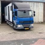 Blue DAF LF cab and white living box pulling out of a warehouse onto brick paving