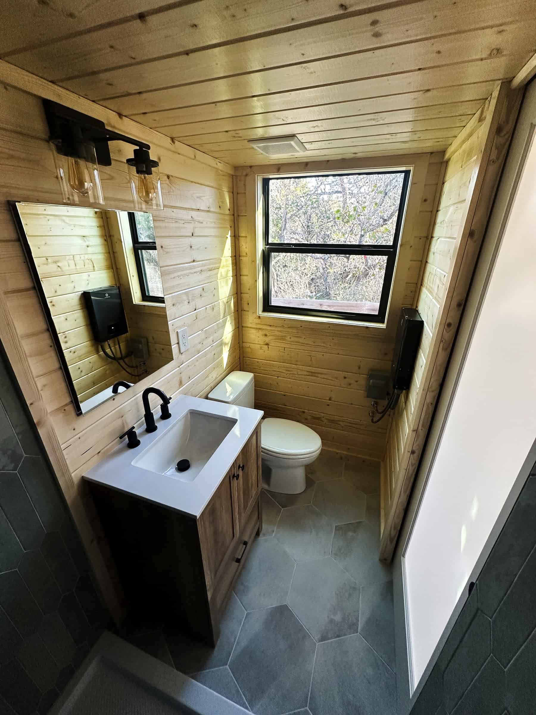 Nomad bathroom: light wood walls, grey hex floor tile, white flush toilet under a black-framed window, dark wood vanity with a white sink