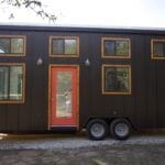 Charcoal vertical-siding Nomad tiny house on a dual-axle trailer on gravel: coral-red glass door, light wood trim, four clerestory windows, Dripping Springs Texas
