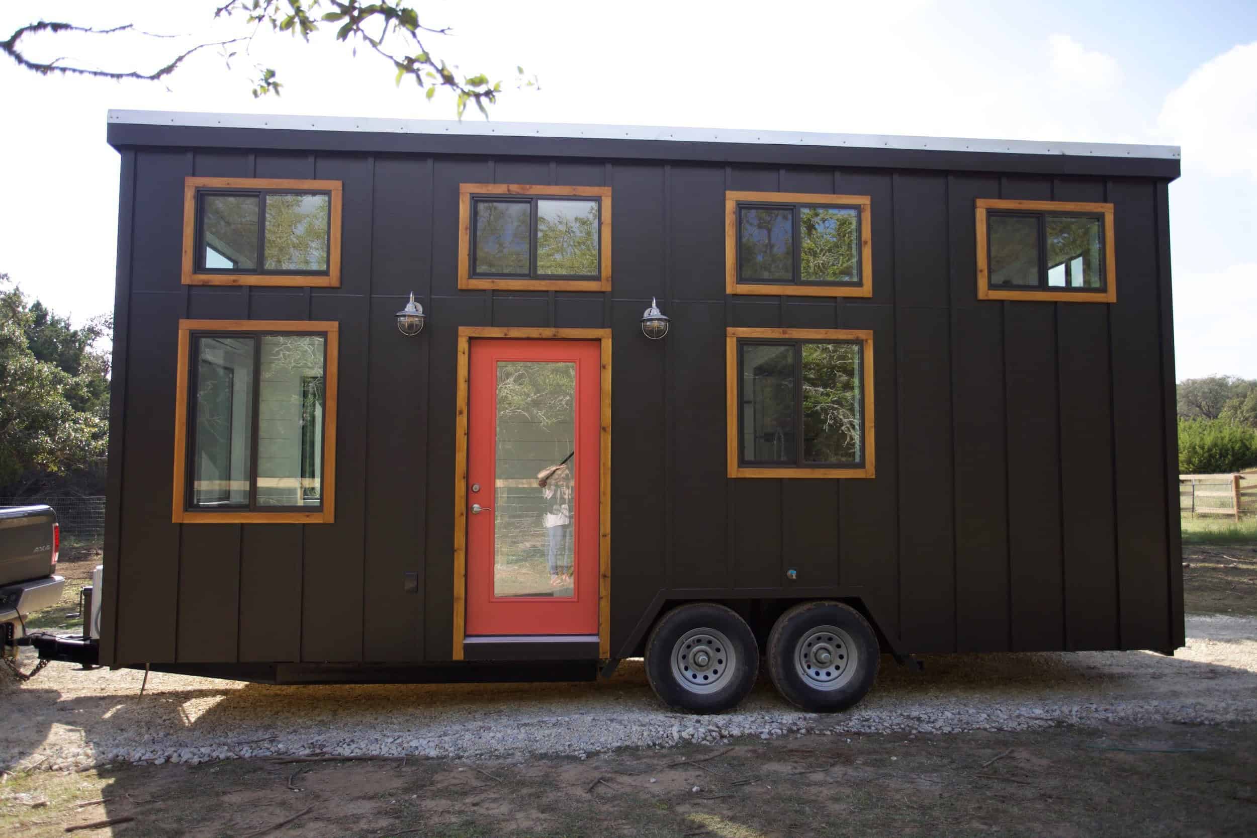 Charcoal vertical-siding Nomad tiny house on a dual-axle trailer on gravel: coral-red glass door, light wood trim, four clerestory windows, Dripping Springs Texas