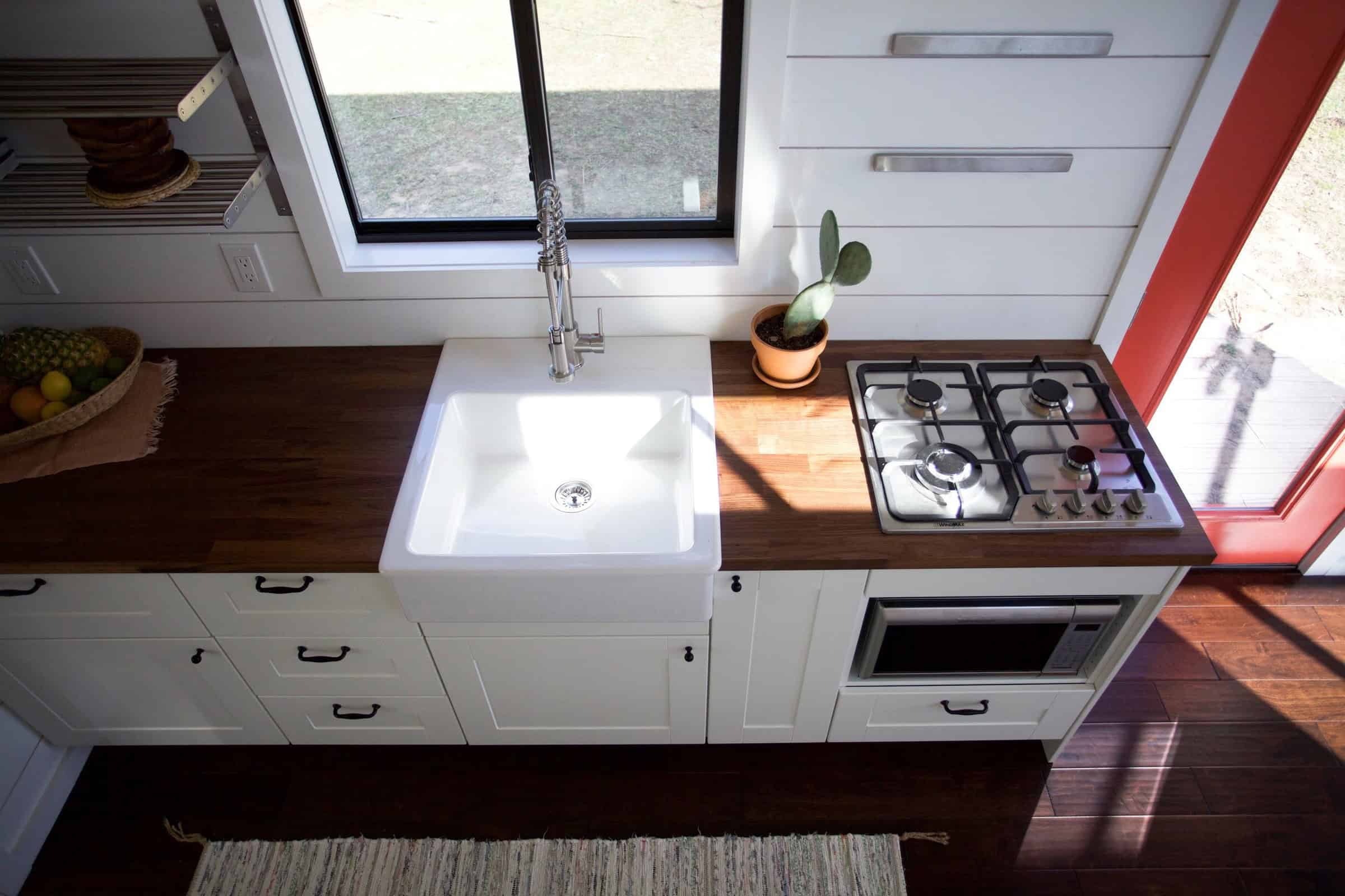 Nomad kitchen: white farmhouse sink, butcher-block counter, cooktop, white shaker cabinets, black-framed window, coral-red door frame at the right