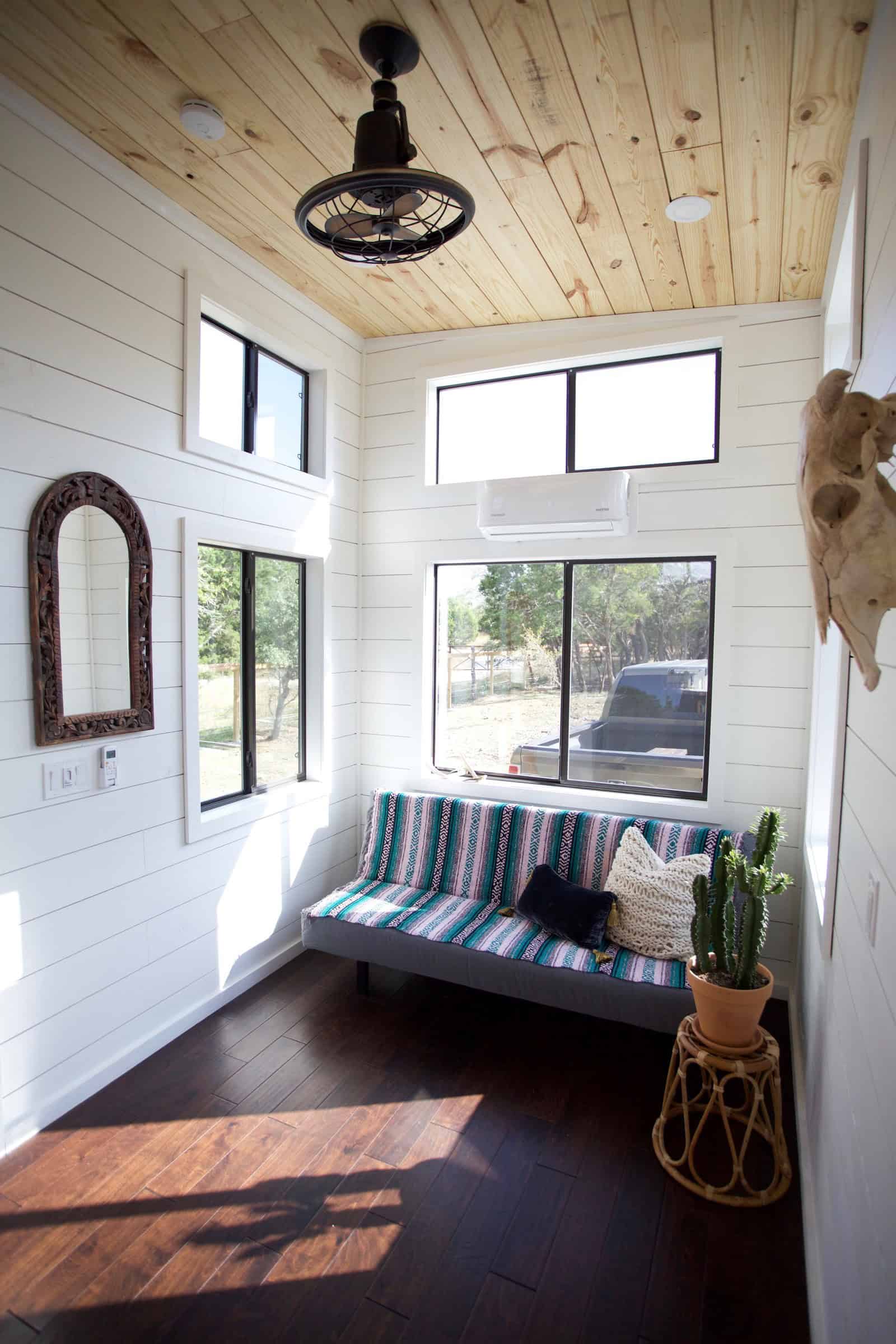 Nomad living area: white shiplap walls, wood plank ceiling, grey sofa with a striped throw, black-framed windows, dark wood floor