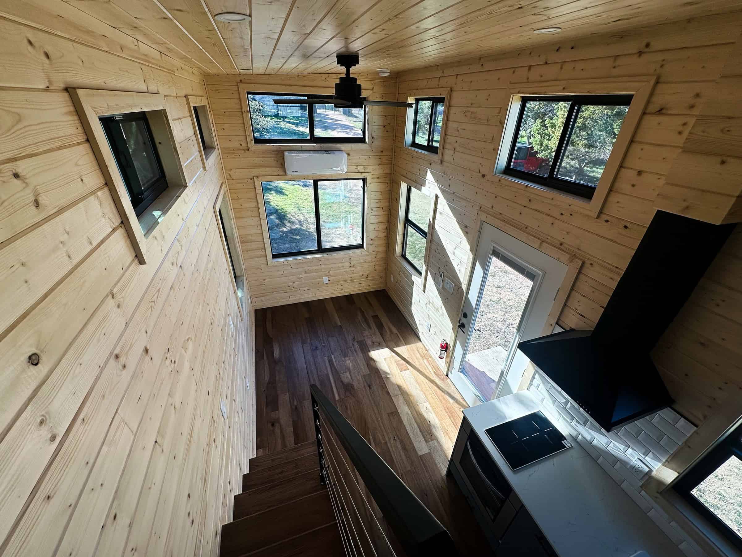 Looking down from a Nomad loft: wood tongue-and-groove walls and ceiling, cable stair rail, kitchen cooktop and black range hood, glass entry door