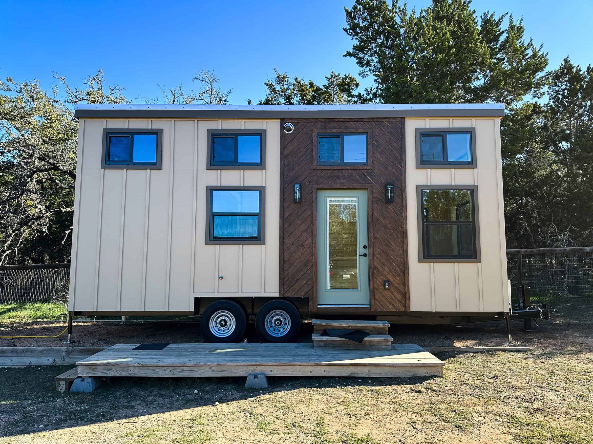 Tan board-and-batten Nomad tiny house on a dual-axle trailer: chevron wood around a sage-green glass door, small wood deck, four clerestory windows