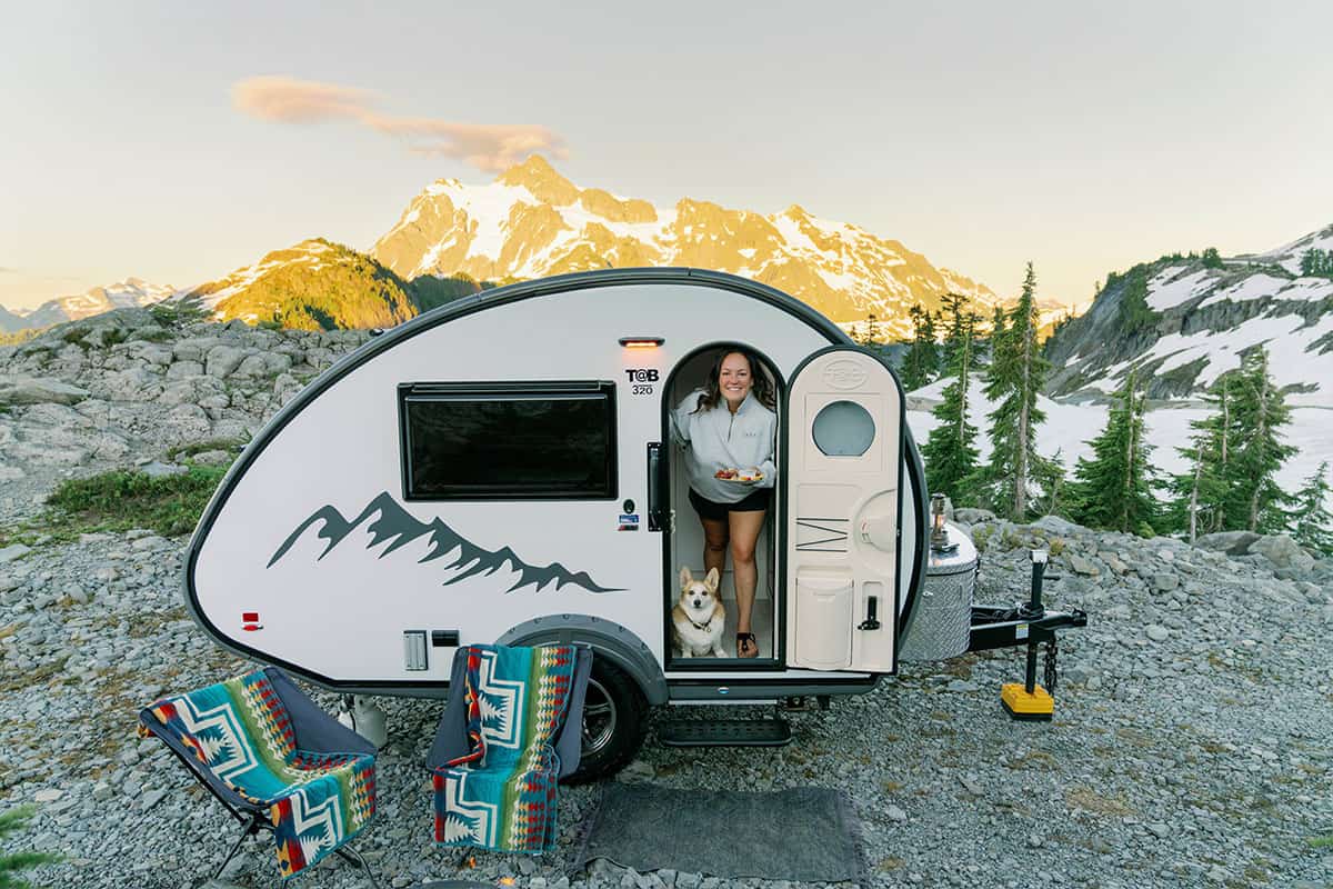 2026 nuCamp TAB 320 teardrop with the side door open on a rocky ridge, person and Corgi in the doorway, camp chairs in the foreground