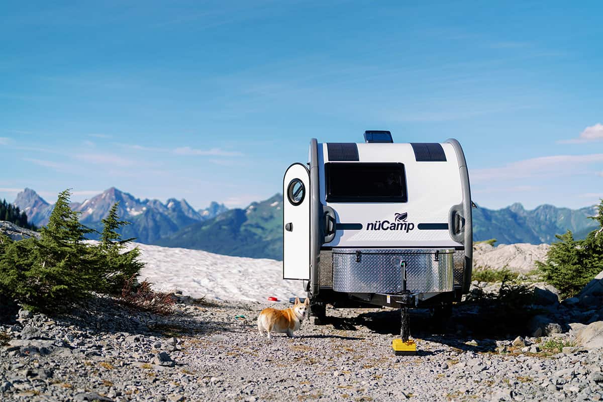 2026 nuCamp TAB 320 teardrop from the rear on a rocky path: two solar panels on the roof, door open, Corgi on the gravel