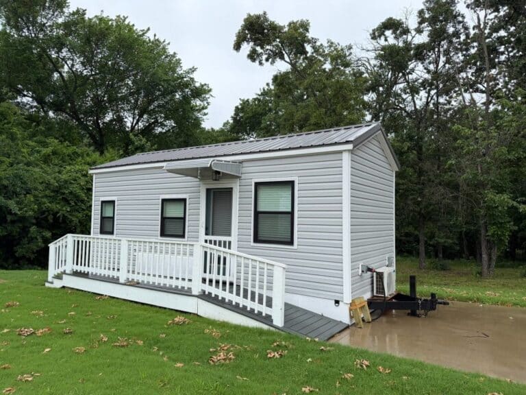 Grey tiny house on wheels with a porch, ramp, and trailer tongue on a concrete pad at The Oaks on 37 in Mount Vernon, Texas