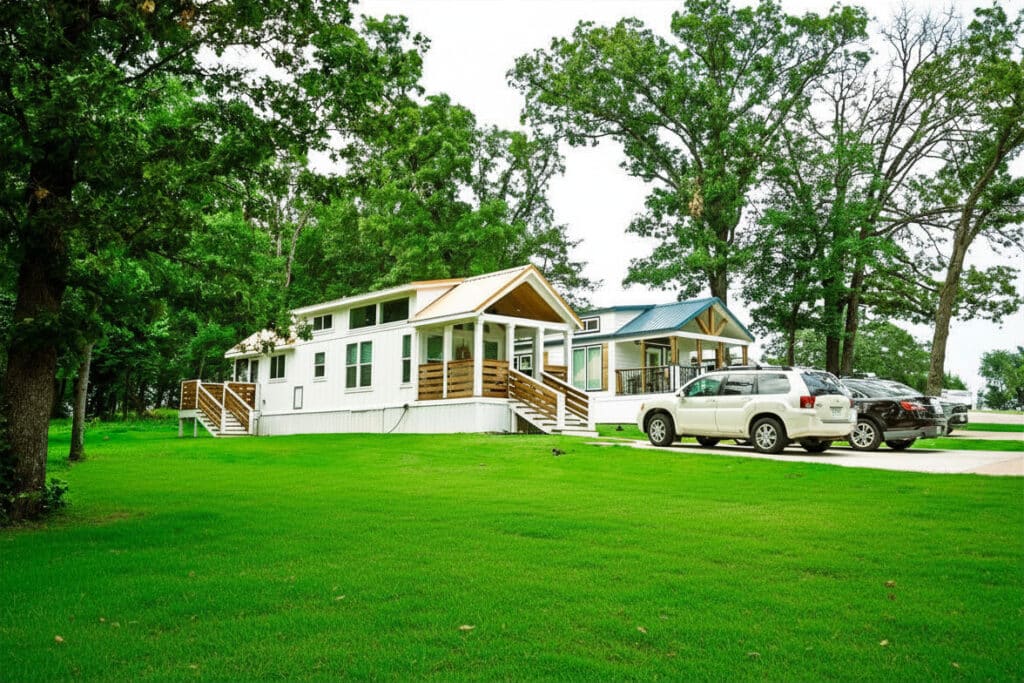 Two white tiny-home cottages on a green lawn with cars parked on concrete pads at The Oaks on 37 in Mount Vernon, Texas