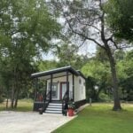 White tiny home with black trim, a red front door, a porch, and a concrete parking pad at The Oaks on 37 in Mount Vernon, Texas