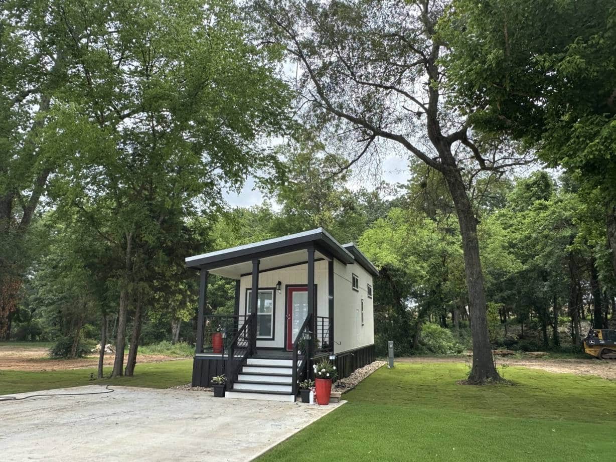 White tiny home with black trim, a red front door, a porch, and a concrete parking pad at The Oaks on 37 in Mount Vernon, Texas