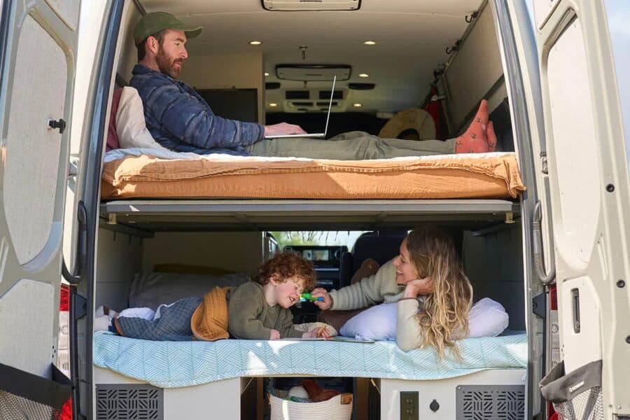 Rear of the Approach through open barn doors: upper three-panel bed with terracotta bedding and a man on a laptop, lower bench-bed with a woman and child, garage basket and outlets below