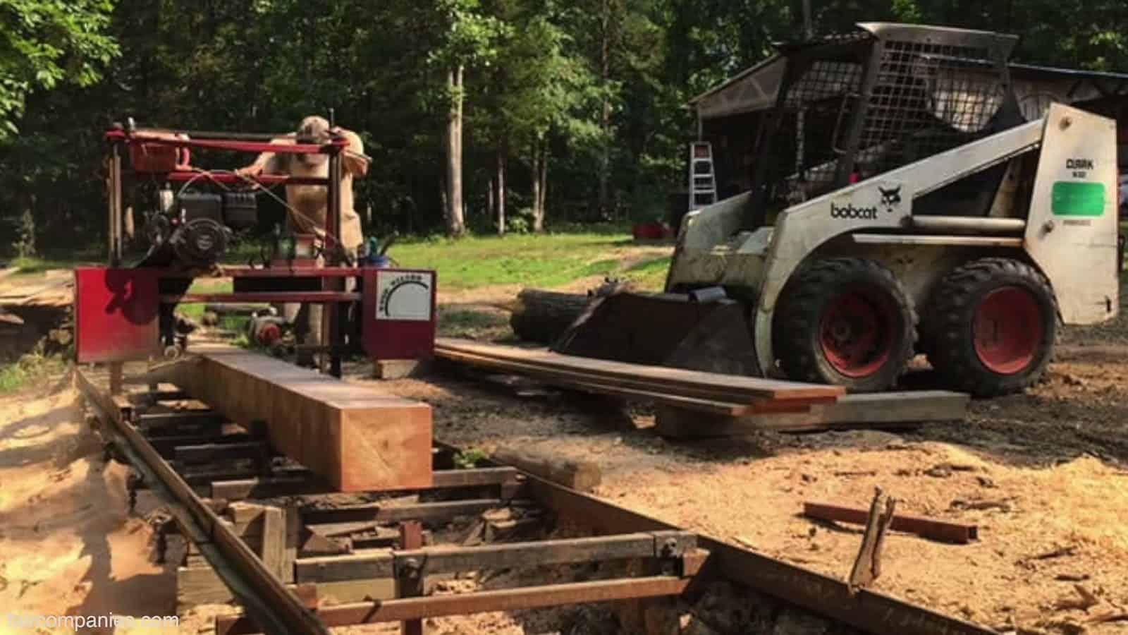 Man running a portable band sawmill beside a freshly milled timber, with a skid steer parked in a clearing in the Ozarks