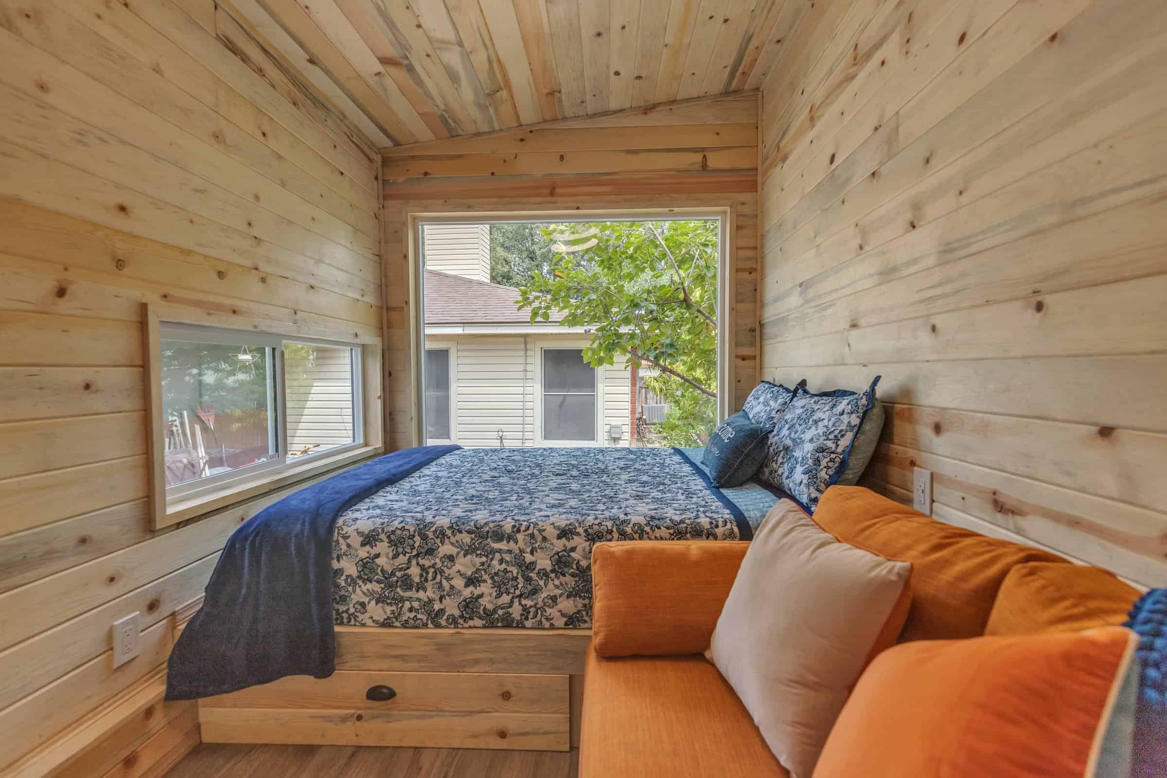 Pacifica Model X studio bed and lounge: platform bed with floral quilt, under-bed drawer, orange sofa, vaulted wood tongue-and-groove