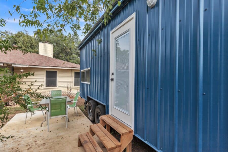 Pacifica Model X exterior: deep blue corrugated metal siding, white trim, full-view glass door, wood steps, dual-axle trailer beside a patio