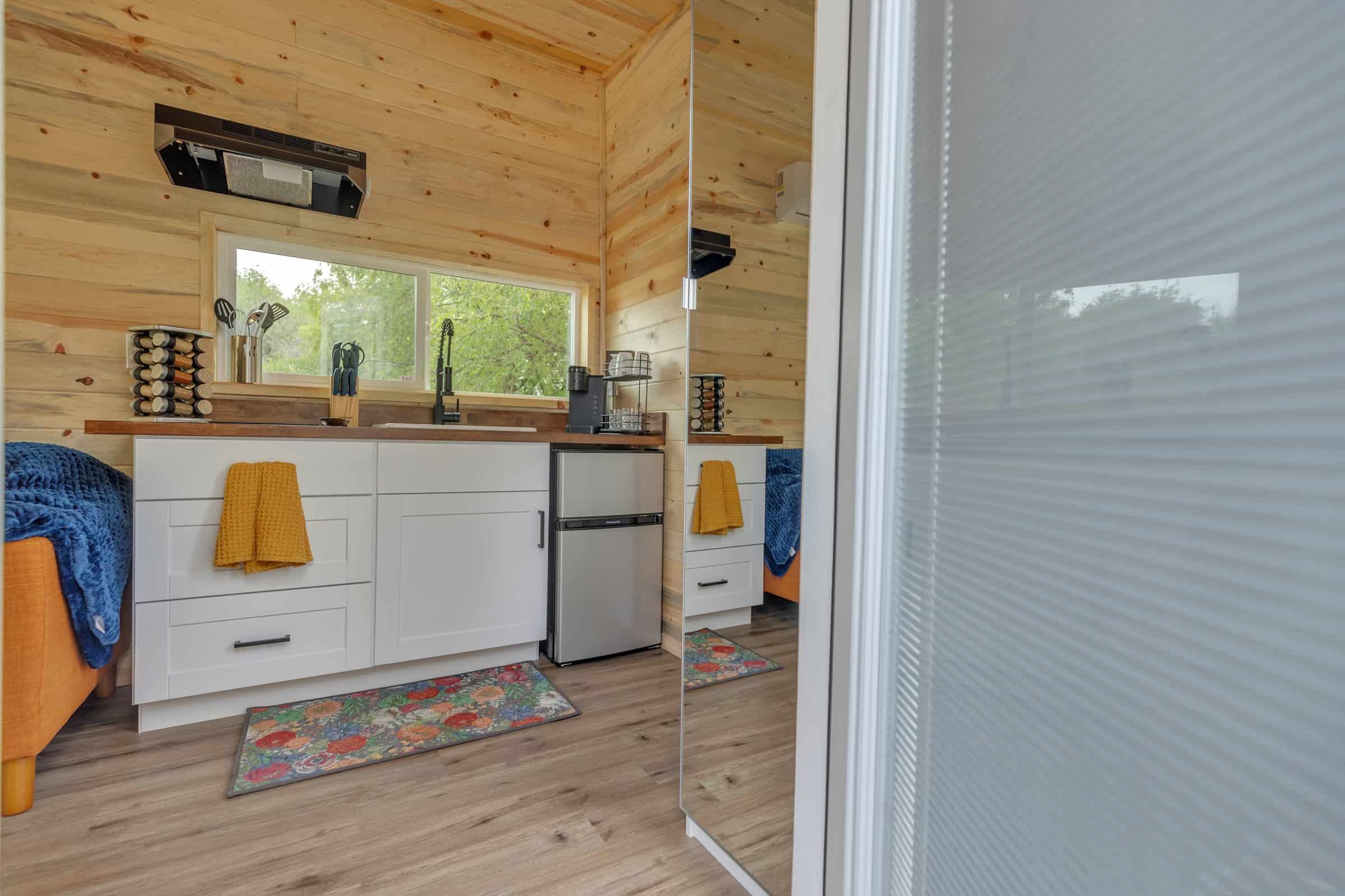 Pacifica Model X kitchen: light wood tongue-and-groove walls and ceiling, white cabinets, butcher-block counter, black faucet, mini-fridge, orange sofa edge