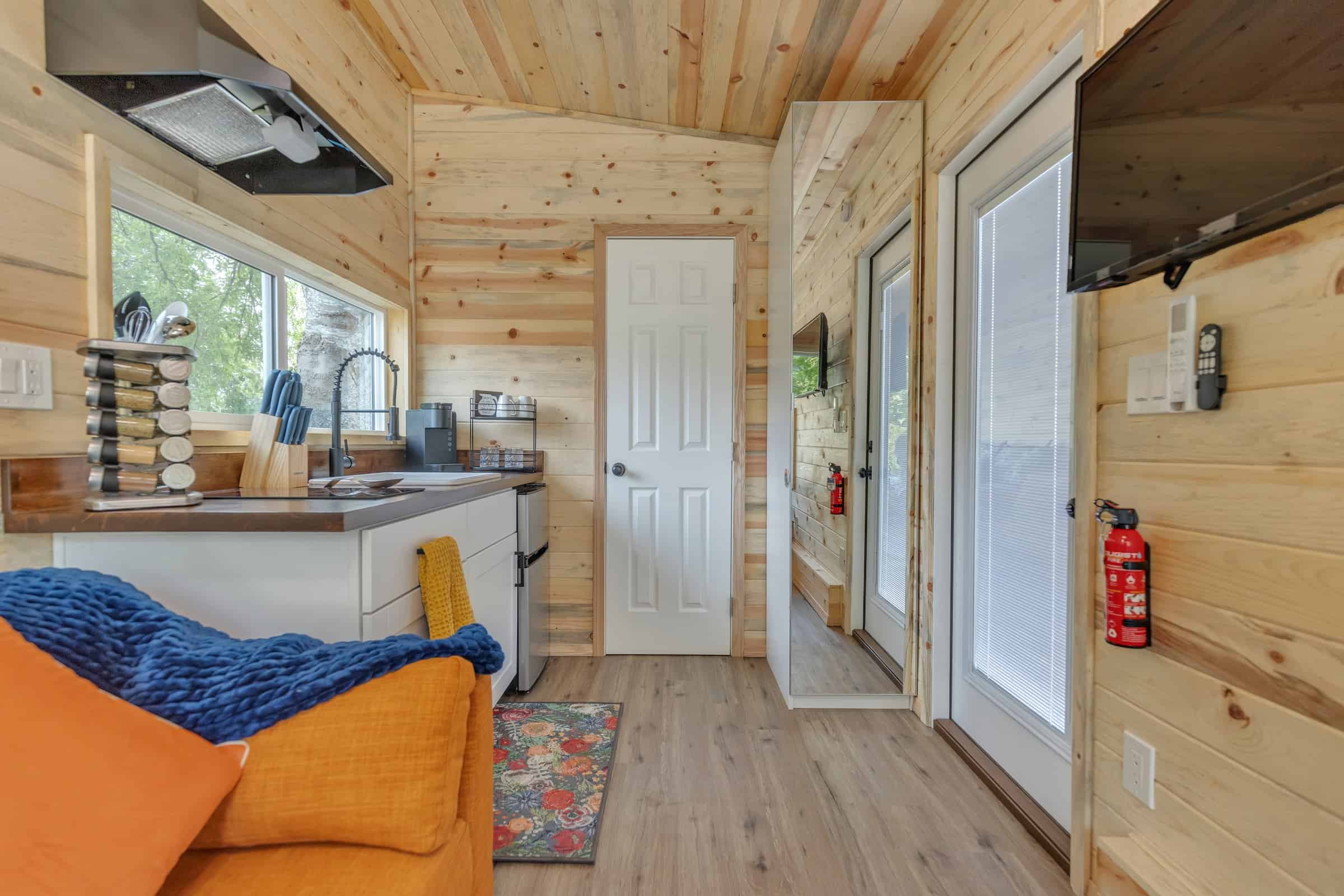 Pacifica Model X living toward kitchen and entry: orange sofa, white cabinets, butcher-block, mirrored wardrobe, glass door, TV on the wall