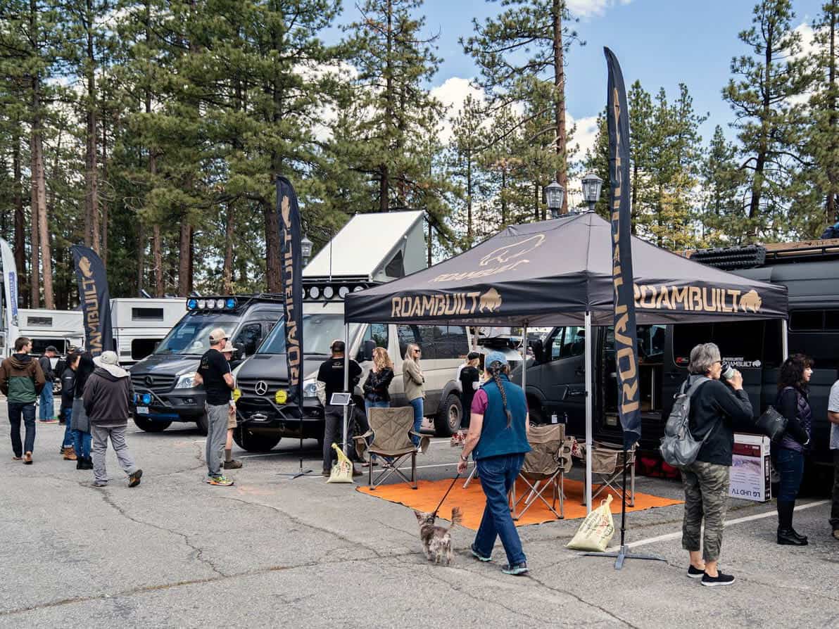 Roambuilt booth and adventure vans on the pavement at Palisades Tahoe, pine trees and Olympic Valley behind them