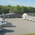 Aerial of the Phillips Happy Hallow sales-center lot in Lakeview, Ohio — park-model houses lined on the right, fifth-wheel RVs on the left, a dark metal building with a red awning, gravel lot and woods. This is the commercial sales lot, not the Park Model Paradise HOA streets.