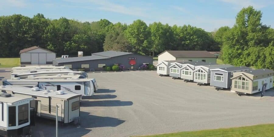 Aerial of the Phillips Happy Hallow sales-center lot in Lakeview, Ohio — park-model houses lined on the right, fifth-wheel RVs on the left, a dark metal building with a red awning, gravel lot and woods. This is the commercial sales lot, not the Park Model Paradise HOA streets.