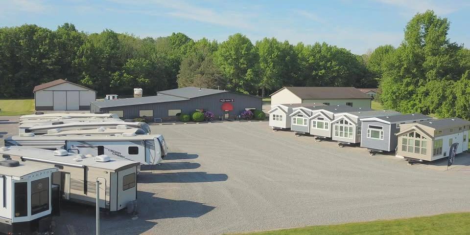 Aerial of the Phillips Happy Hallow sales-center lot in Lakeview, Ohio — park-model houses lined on the right, fifth-wheel RVs on the left, a dark metal building with a red awning, gravel lot and woods. This is the commercial sales lot, not the Park Model Paradise HOA streets.