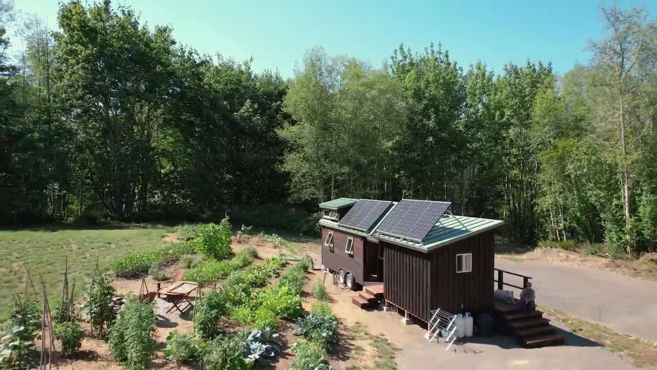 Aerial view of a shou sugi ban tiny house and matching workshop joined by a cedar deck, solar arrays on both roofs, garden, Patrick and husky Barley
