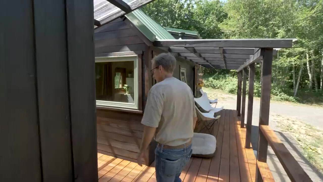 Covered cedar deck with a translucent roof, butterfly chairs, and a dog bed between the tiny house and workshop