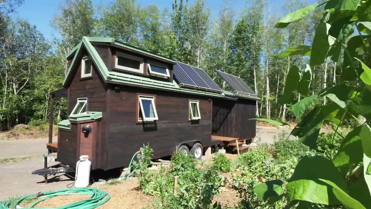 Side elevation of a dark shou sugi ban redwood tiny house on a dual-axle trailer with loft dormers and a green metal roof