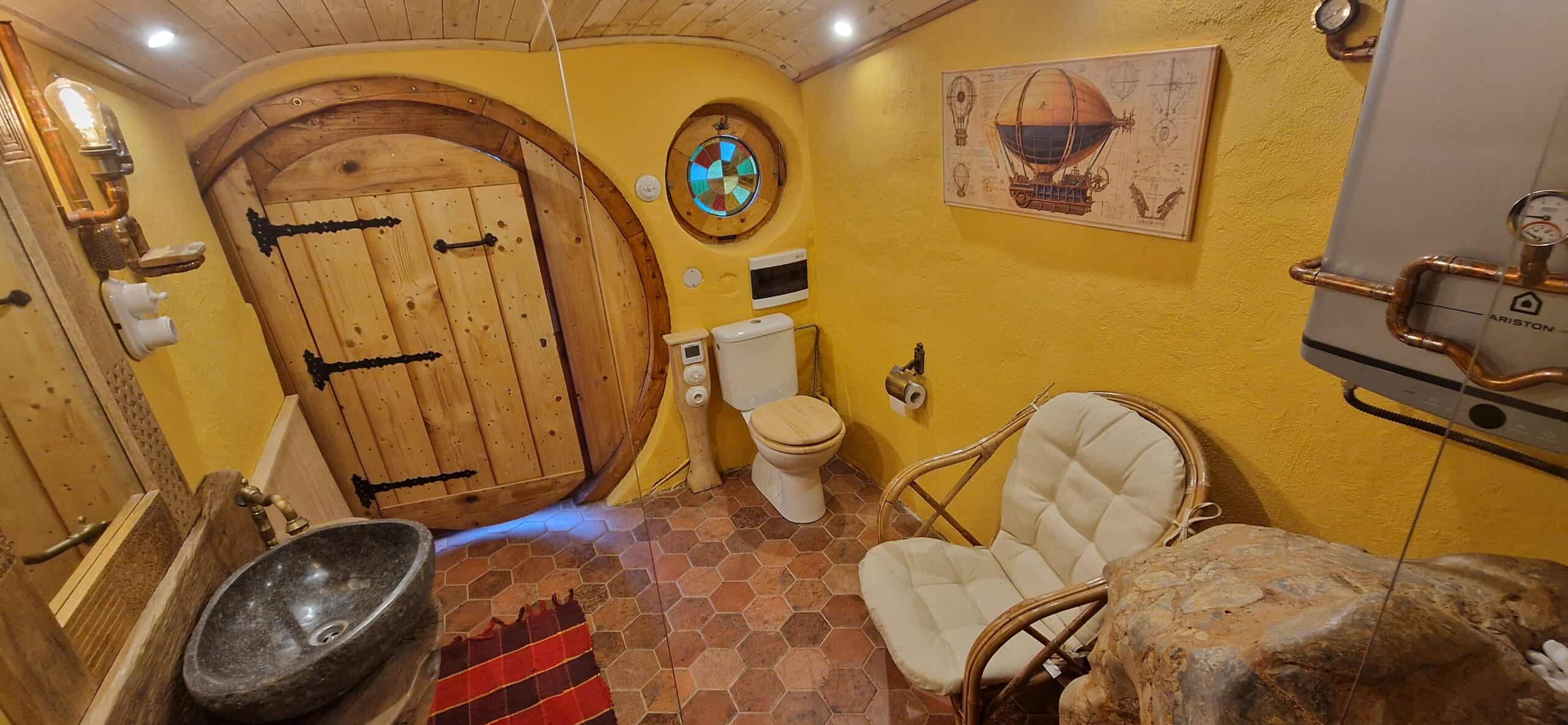 Hobbit bathroom interior with yellow walls, round wooden door, stone vessel sink, and terracotta tile floor