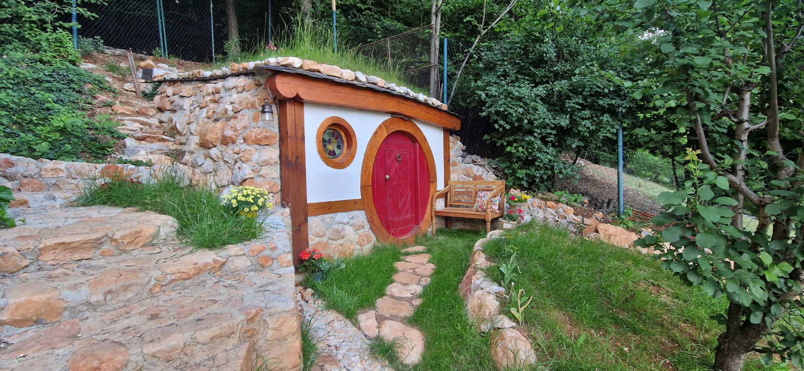 Hobbit-style hillside bathroom building with round red door, stained-glass window, and living grass roof