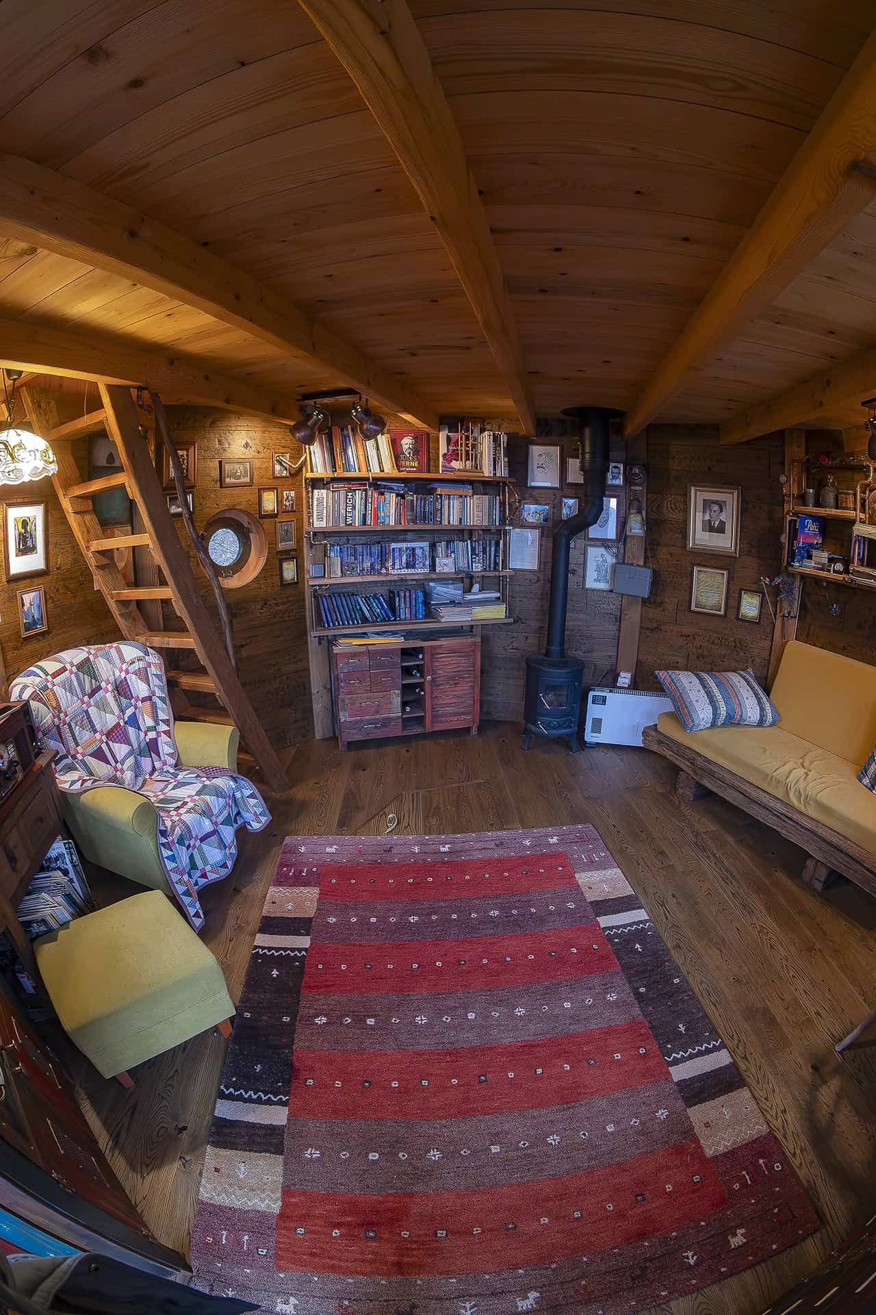 Cozy wooden living area inside the tower with bookshelf, wood stove, yellow armchair, and ladder to the bedroom