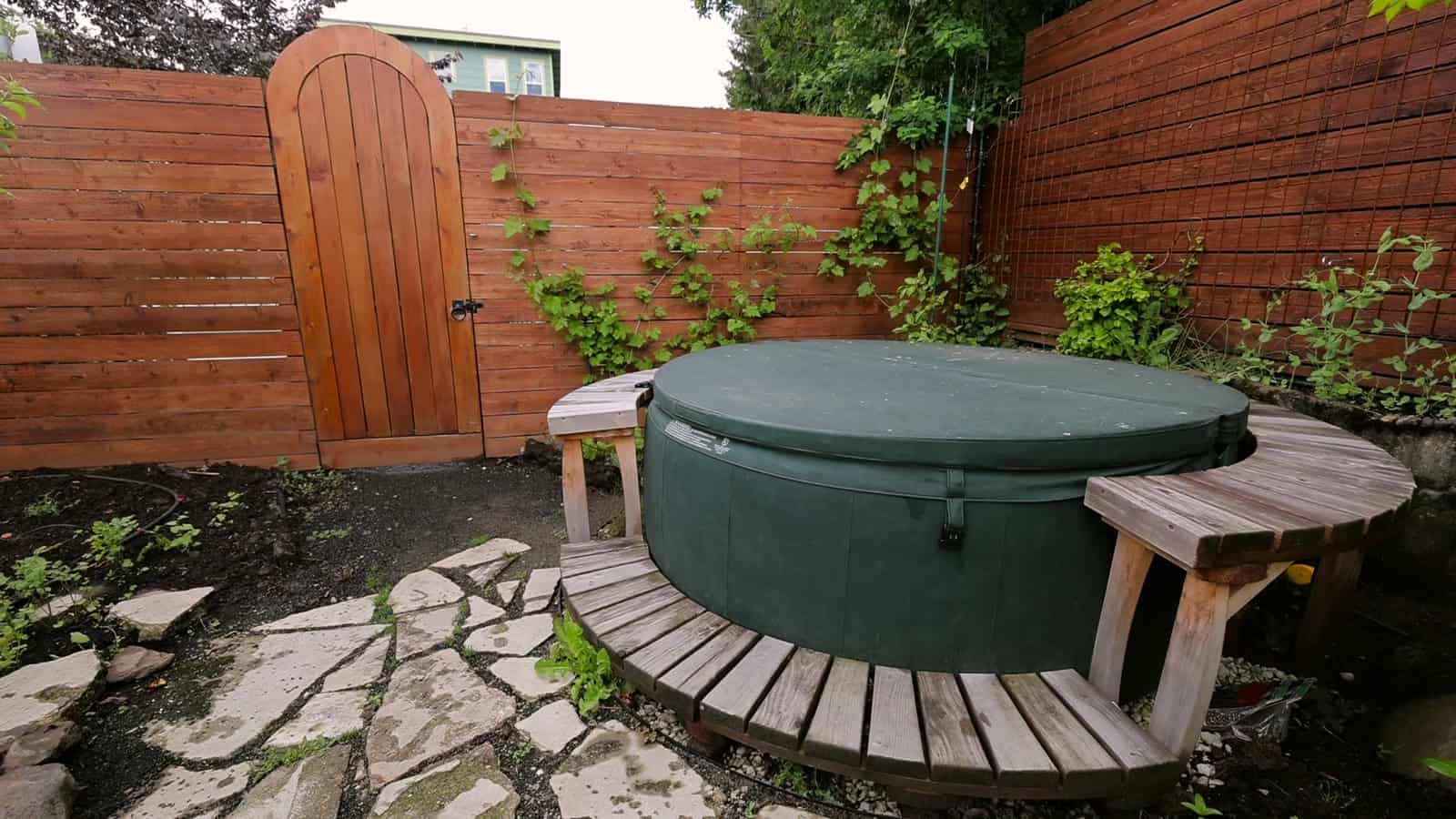 Round green hot tub with curved wooden bench seating beside an arched wooden gate and flagstone path in a Portland backyard