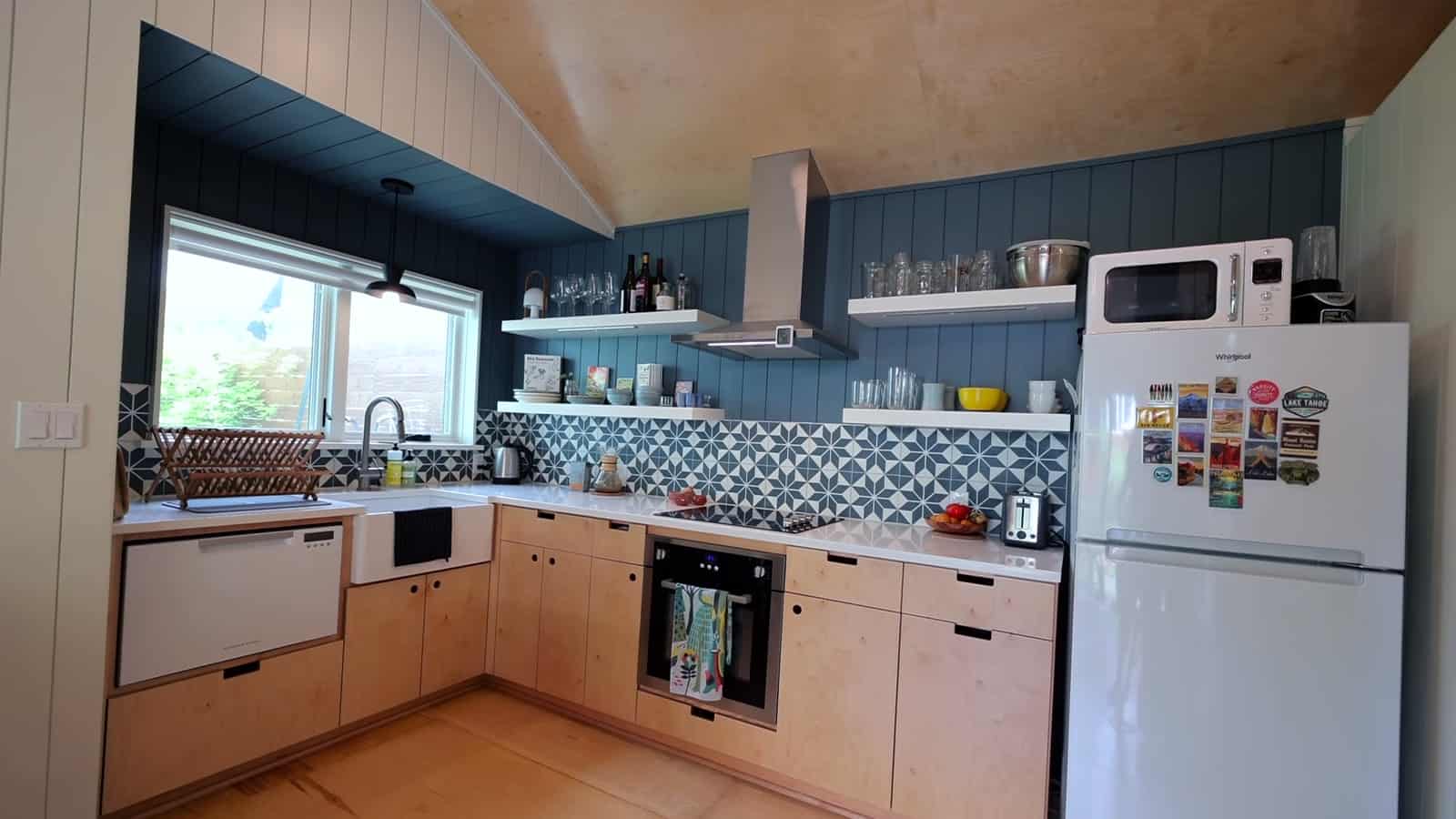 Kitchen with Baltic birch plywood cabinets, blue-gray vertical paneling, patterned tile backsplash, open shelves, and white refrigerator