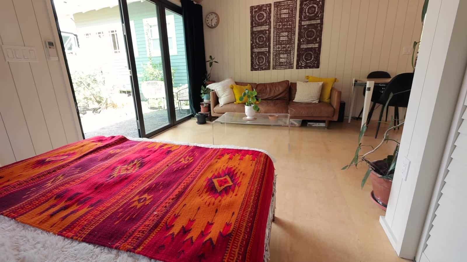 Studio interior looking past a bed with a bright red patterned throw toward an open panoramic door, leather sofa, and garden beyond