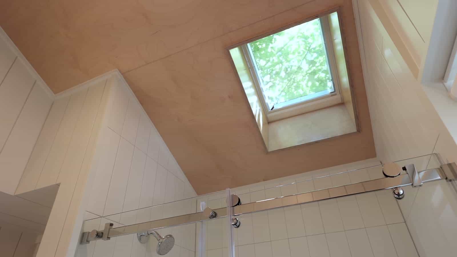 Looking up at an operable skylight set into a wood-lined sloped ceiling above a glass-enclosed tiled shower