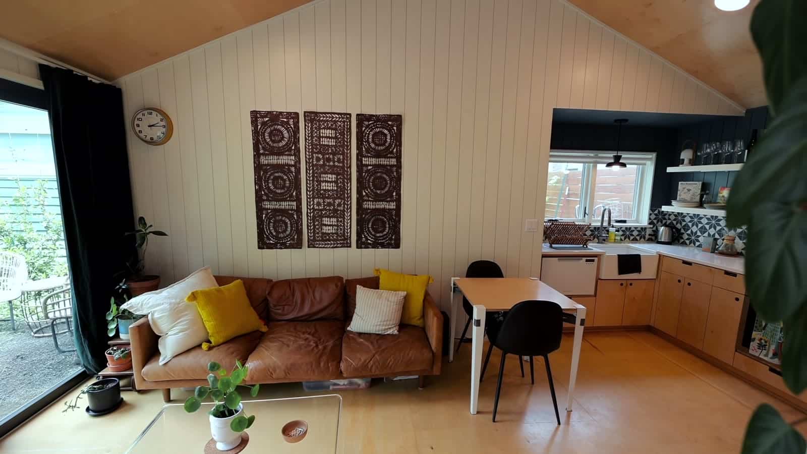Vaulted ceiling studio with leather sofa, framed panels, small dining table, and plywood kitchen in a converted Portland garage ADU
