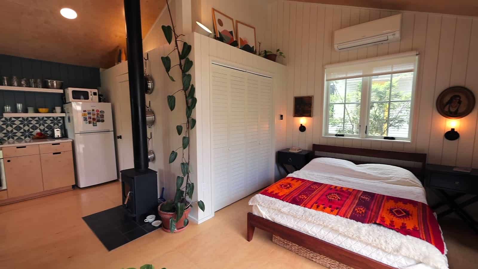 Small black wood stove beside a bedroom nook with a red patterned bedspread, window, wall-mounted TV, and louvered closet doors