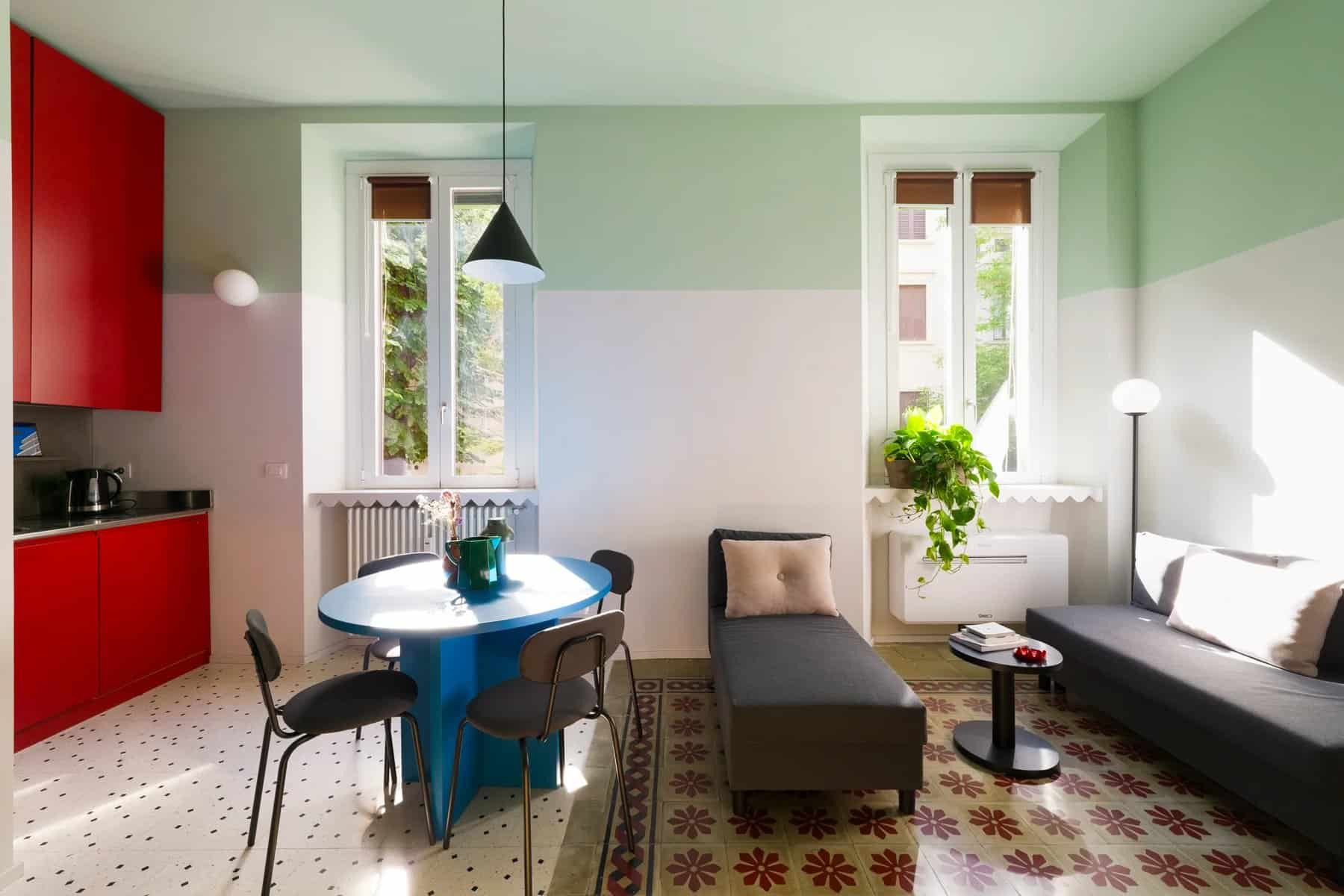 Dining area with a blue round table, black chairs, red kitchen cabinets, and tall windows
