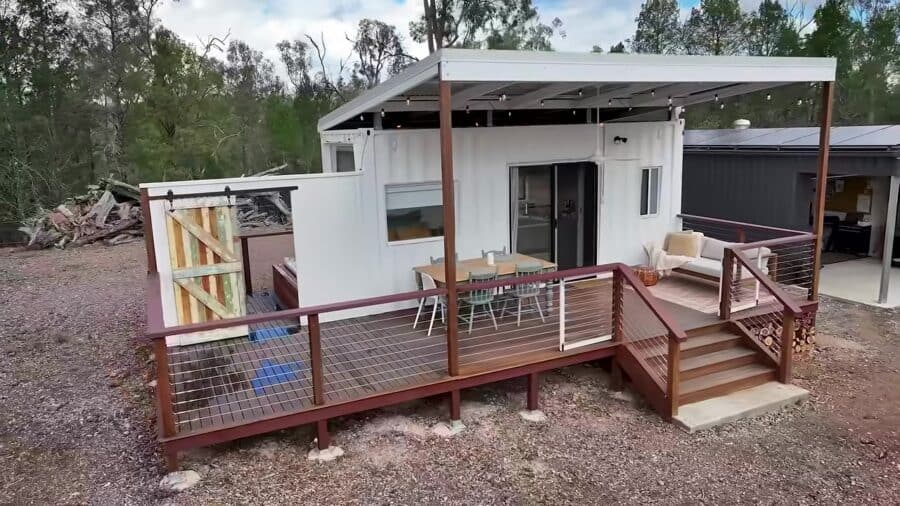 White 20-foot shipping container with a large covered timber deck, dining table, outdoor sofa, and a dark metal shed beyond, on bushland in Queensland