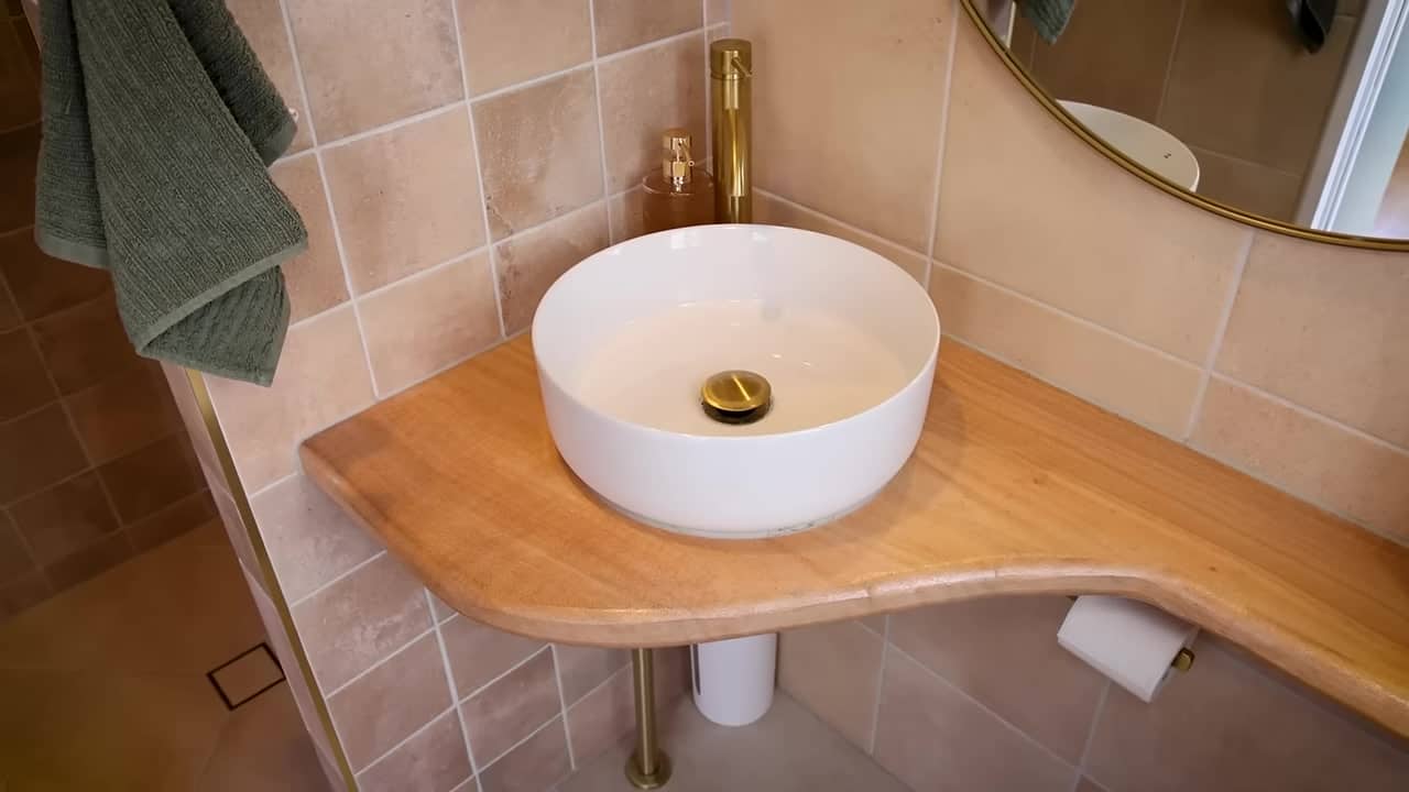 Compact bathroom with a curved live-edge timber vanity, white vessel sink, gold faucet, and sandy natural tiles