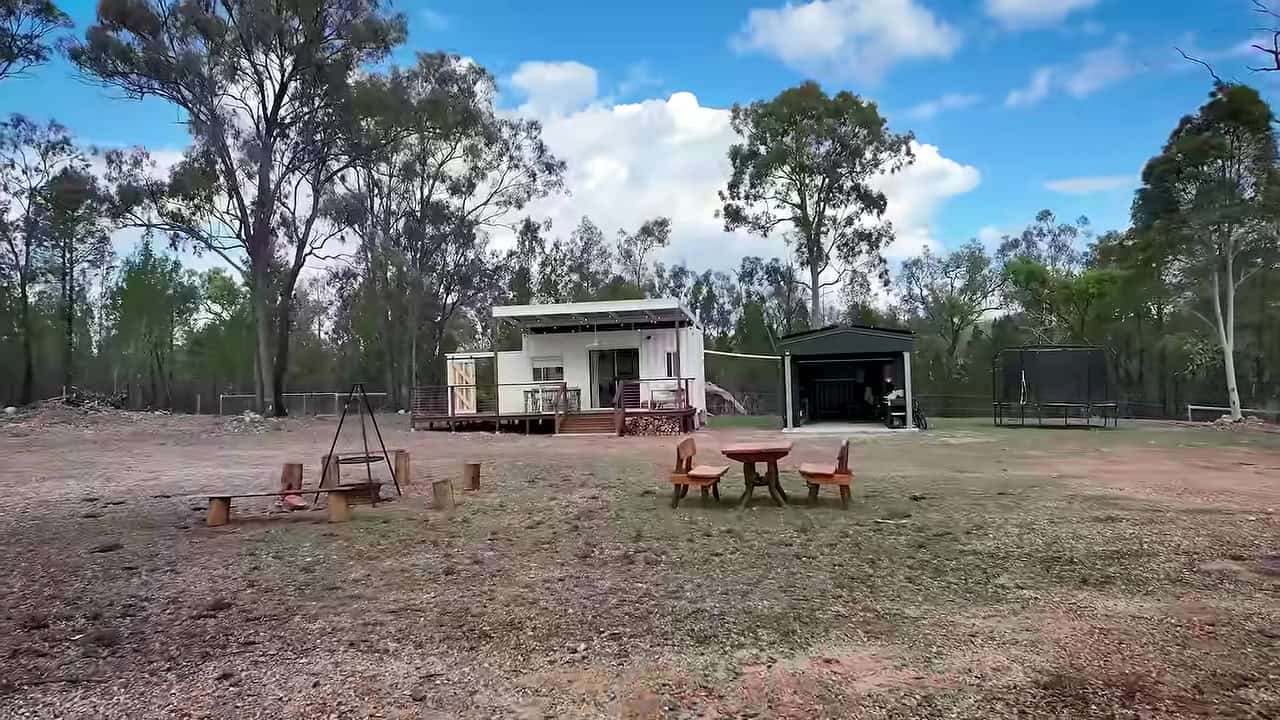 Fire pit with log benches and a cooking tripod in front of a white 20-ft container home and shed on a Queensland property