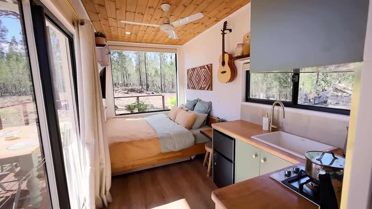 Compact 20-ft container interior looking from the green-and-timber kitchen toward a king bed and a large picture window