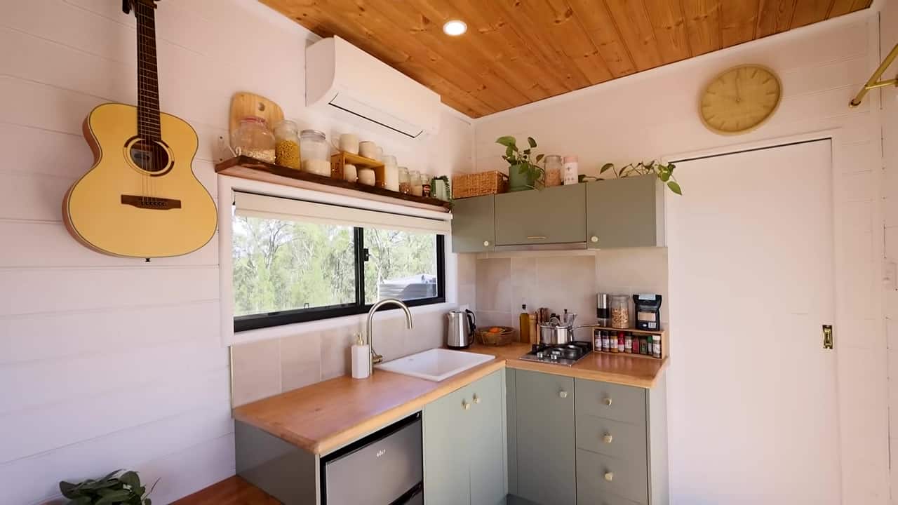 L-shaped kitchen with sage-green cabinets, live-edge timber benchtop, large white sink, gold faucet, two-burner cooktop, and a small fridge